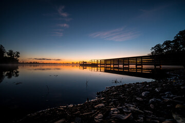 Obraz premium A beautiful sunset over a lake with a wooden bridge in the foreground. The water is calm and the sky is filled with clouds. The scene is peaceful and serene