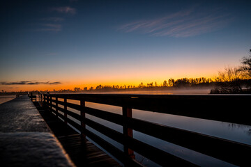 Obraz premium A wooden bridge over a body of water with a beautiful sunset in the background. The water is calm and the sky is filled with clouds