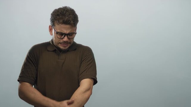 Man wearing brown polo and glasses, arms crossed and tense expression with closed eyes in a pale blue studio; discomfort.