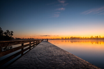 A pier with a dock and a man fishing. The sky is orange and the water is calm