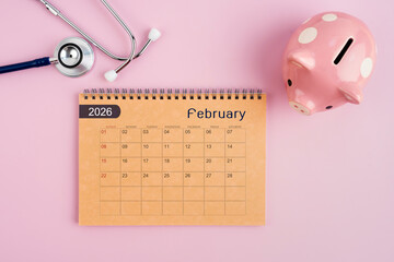February 2026 desk calendar and stethoscope with piggy bank. Savings for Health concept.