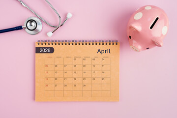 April 2026 desk calendar and stethoscope with piggy bank. Savings for Health concept.