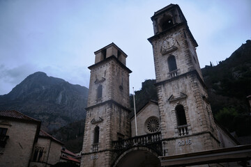 Low-angle view of historic stone cathedral with dual bell towers set against misty mountains on overcast rainy day. The Cathedral of Saint Tryphon in Kotor, Montenegro