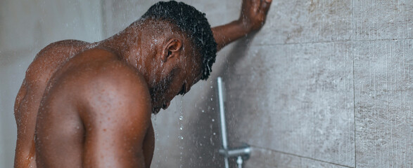 A man showers in his bathroom, water cascades from the showerhead over him. He appears focused on his routine. The setting has modern tiles and a simple design. © Prostock-studio