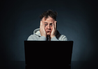 Sad stressed man holding his head while sitting in front of a laptop on dark background. Concept of...