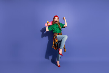 Young woman jumps with a skateboard in colorful casual streetwear against a purple background