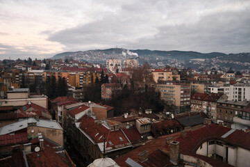 High angle view of densely built European-style city under overcast winter sky. Red rooftops at foreground, apartment blocks extend toward distant hills and mountains. Sarajevo, Bosnia and Herzegovina
