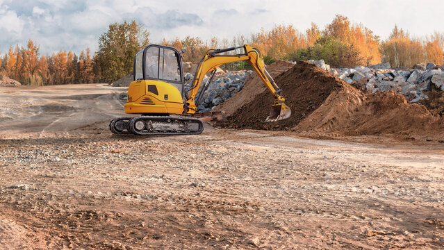 A mini excavator works on a construction site, moving soil and preparing the ground in a residential area in the fall, when the trees are brightly colored