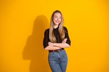 Young woman in casual fashion stands against a bright yellow background smiling with arms folded...