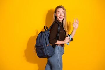Young woman with backpack waving hello in a bright casual fashion style against a bold yellow...