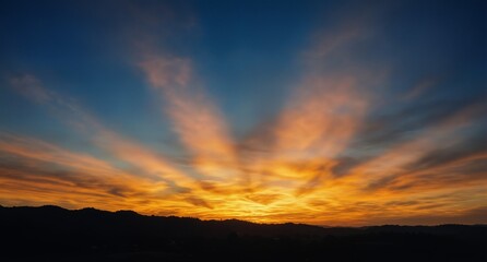 dramatic crepuscular rays burst through vibrant orange and yellow clouds at sunset over silhouetted hills.