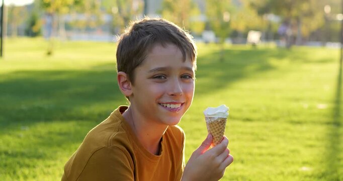 Child smiles in green park and holds ice cream cone at sunset. Child enjoys summer day outdoors. Happy boy relaxed in sunlight. Warm lifestyle scene. childhood joy, leisure time. Sweet food pleasure.
