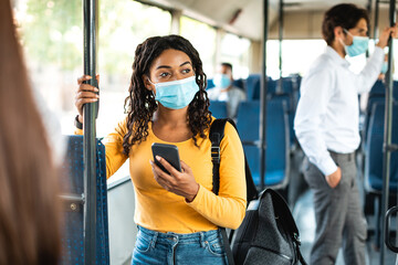 Obraz premium Public Transportation. African American woman in medical mask holding and using smartphone standing in a city bus. Female passenger looking away at window, waiting for her stop, browsing route