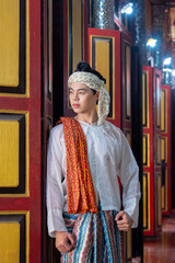 Fototapeta premium Handsome Myanmar man wearing colorful fabric and headscarf standing by ornate wooden doors with warm lighting