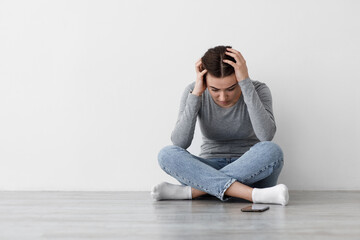 Despaired unhappy sad caucasian millennial female suffering from depression holding head with hand and sitting on floor with smartphone at home. Grief, bad news, self-isolation covid-19 quarantine