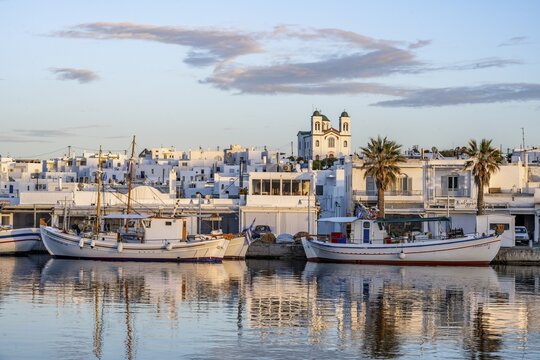View of Naoussa, Fishing boats in the harbour at sunset, reflected in the sea, White Cycladic houses and church Agios Faneromeni, Naoussa, Paros, Cyclades, Greece