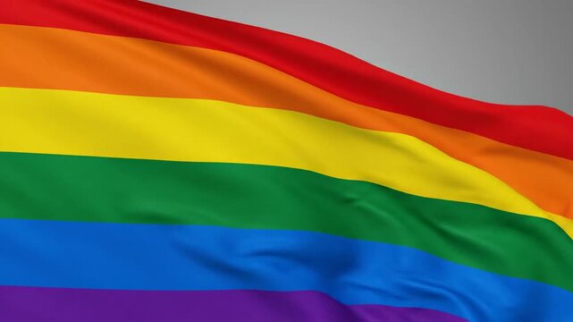 The iconic rainbow LGBTQ pride flag fluttering in the wind against a clear blue sky with a few scattered clouds.