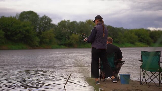 Young couple man and woman fishing on river bank on cloudy day. Spending time together. Relaxing family holidays by the river.