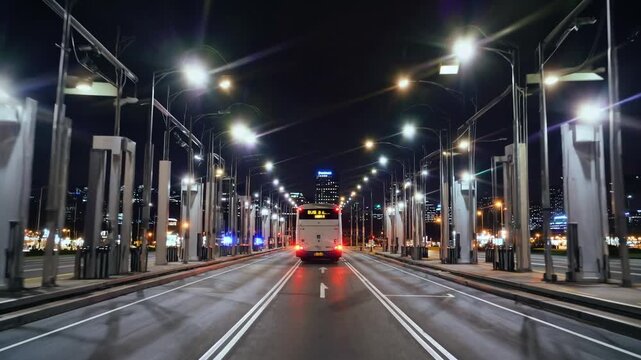 POV shot driving through a brightly lit, empty highway toll plaza at night. A white bus is proceeding through the automated payment gate