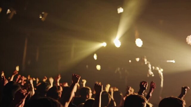 Back view of a large crowd of young people dancing with their hands up at a music festival. A singer performs on a stage lit by spotlights