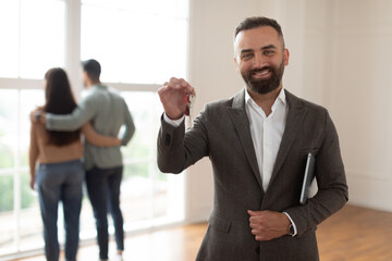 Portrait Of Cheerful Professional Real Estate Agent Holding Clipboard And Showing Keys In Hand, Selective Focus. Young Newlyweds Choosing New Home Looking Out Window Hugging In Blurred Background
