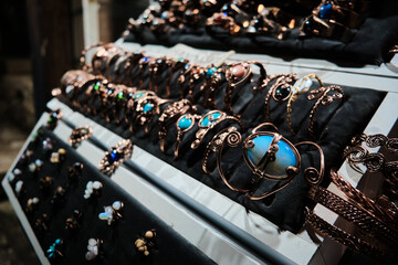 Close-up view of handcrafted copper bracelets and rings with turquoise and gemstone details arranged on a jewelry stand. Artisanal metalwork and polished stones create rustic market aesthetic