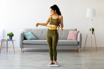 Full length of pretty Indian woman measuring her waist while standing on scales, having perfect body parameters at home. Young Eastern lady satisfied with result of her slimming diet, free space © Prostock-studio
