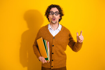 Young man with glasses wearing a brown cardigan holds folders against a bright yellow background and points with confidence
