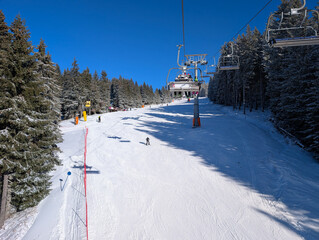 Chairlift with riders above wide ski slope in Kopaonik forest. Winter mobility, alpine infrastructure, and recreational tourism through transport, snow covered terrain, and mountain leisure culture.