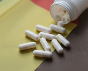 Several white medication capsules are shown spilled from their white plastic container onto a yellow and brown surface.