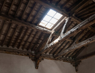 The weathered wooden truss structure of an old roof features exposed beams and rafters illuminated by a central skylight.