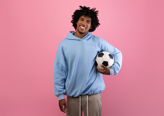 Portrait of positive black teenage guy holding soccer ball on pink studio background. Cheerful...