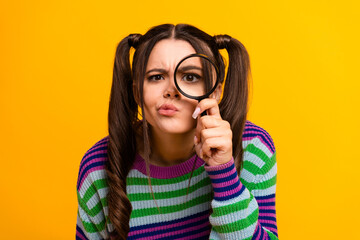 Young woman with striped sweater looks through a magnifying glass against a bright yellow...