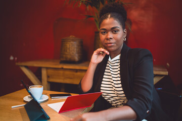 Naklejka premium Black woman poses confidently with tablet, red notebook, and coffee. A portrait of modern freelance work, blending creativity, clarity, and professional digital tools.