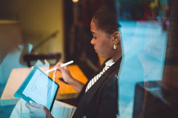 Freelancer draws digital charts with stylus on tablet, working remotely in a café. The window reflection blends her presence with the tools of modern mobile productivity.