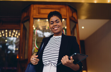 Confident Black woman stands in front of illuminated vintage building holding tablet and stylus,...