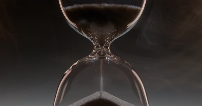 Closeup of hourglass measuring time over a dark dramatic background. Macro view of sand falling down inside the glass clock counting minutes. Concept of time and life flowing.