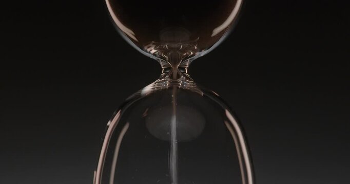 Closeup of hourglass measuring time over a dark dramatic background. Macro view of sand falling down inside the glass clock counting minutes. Concept of time and life flowing.