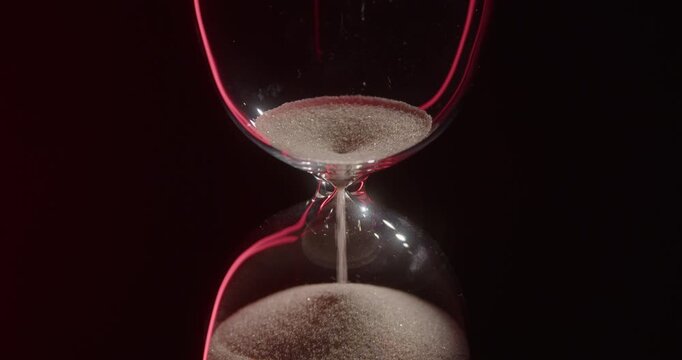 Closeup of hourglass measuring time over a dark dramatic background. Macro view of sand falling down inside the glass clock counting minutes. Concept of time and life flowing.