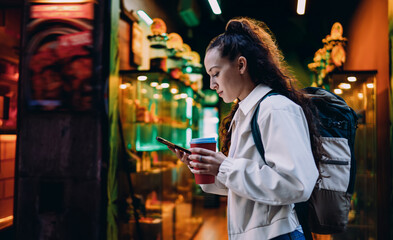 Fototapeta premium Modern woman in casual outfit uses smartphone while walking, coffee in hand, deeply engaged in mobile screen surrounded by glowing shop lights and digital ambiance.