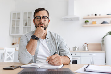 Fototapeta premium Thoughtful middle aged caucasian man student with beard in glasses works at laptop and watch online lesson in minimalist kitchen interior. Brainstorm, creative idea for remote work, study due covid-19