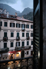 View from open balcony window overlooking historic European-style building with green shutters and balconies. Misty mountains in the background. Old town of Kotor, Montenegro