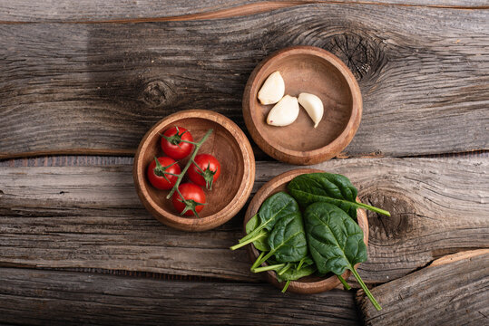 Spinach leaves, cherry tomatoes, and garlic in wooden bowls on wooden table. Top view. Nobody.
