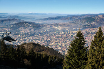 High-altitude perspective looking down over sprawling city nestled between layered mountain ridges. Evergreen trees frame the foreground. Sarajevo, Bosnia and Herzegovina