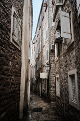 Slender pedestrian alley lined with aged stone buildings leads toward bright opening beneath overcast sky. Weathered masonry, shuttered windows and hanging lanterns. Old town of Budva, Montenegro