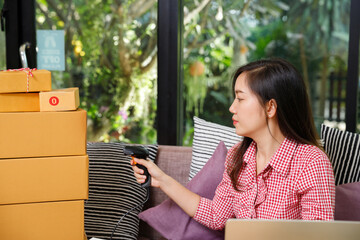 Young Asian SME owner scanning barcode on parcel box for delivery. E-commerce woman checking stock inventory at home office. Logistics and online selling process concept.