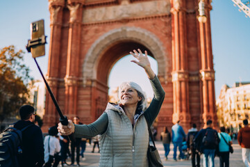 Obraz premium Older woman waves cheerfully while filming selfie video with smartphone and selfie stick. A lively scene showing age diversity and natural confidence with mobile tech.