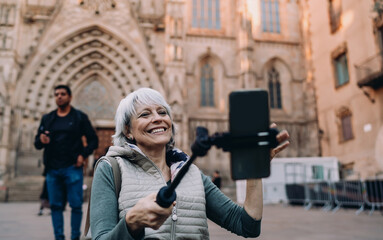 Fototapeta premium Happy senior woman recording herself on smartphone with selfie stick in front of historical cathedral. Captures joyful solo travel, digital expression and confident aging.