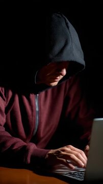 A shadowy figure in a hoodie focused on a laptop in darkness.