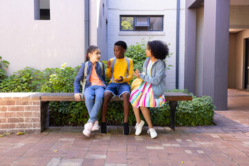 Diverse teenage friends sitting on wooden bench in courtyard, holding backpacks and handheld items